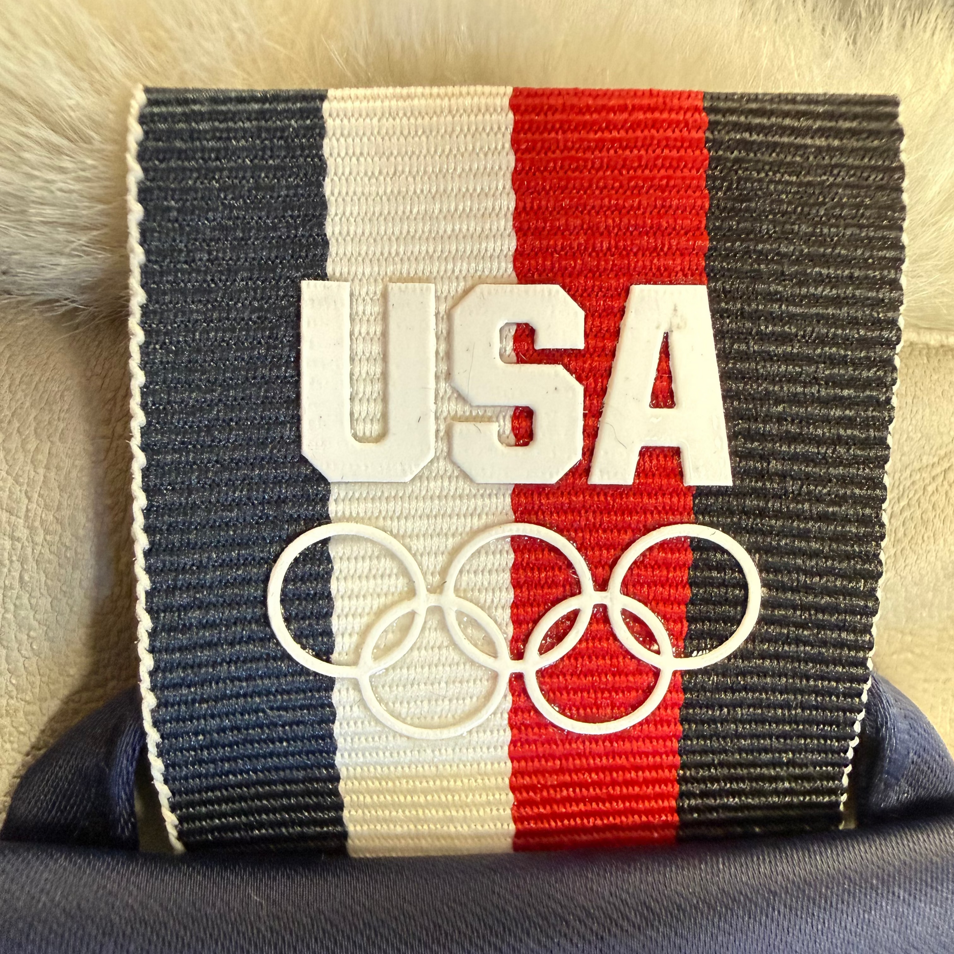 A close-up of a USA Olympic-themed patch or label featuring vertical navy, white, and red stripes, large white 'USA' lettering, and the five Olympic rings on a dark fabric background.