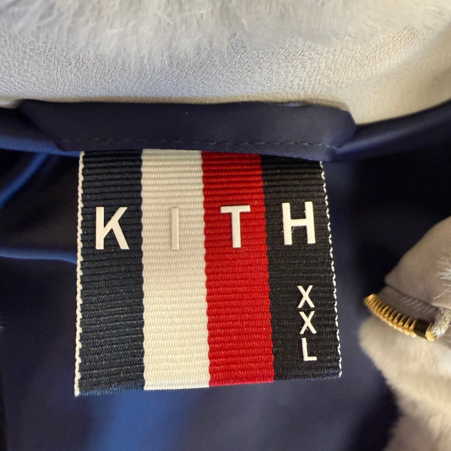 Close-up of a Kith brand fabric label sewn onto a navy blue garment, featuring the iconic red, white, and navy blue vertical stripes with 'KITH' and 'XXL' text, next to a light gray faux fur lining.
